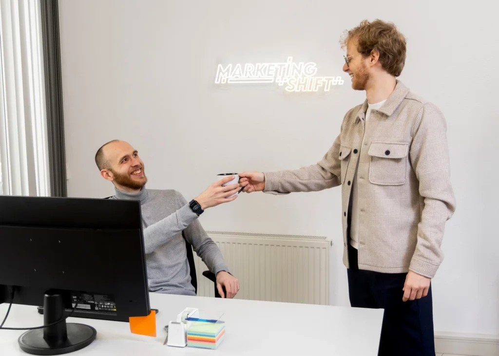 Daniel en Roy bij de balie op kantoor, met het neonlogo van Marketing Shift op de muur